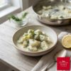 A rustic ceramic bowl filled with creamy, dill-flecked stewed cucumber chunks sits on a weathered wooden table, accompanied by a spoon and lemon.
