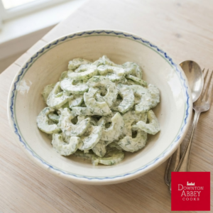 A rustic, patterned ceramic bowl filled with a creamy cucumber and dill salad sits on a light wooden table beside a vintage silver spoon and fork.