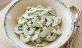 A rustic, patterned ceramic bowl filled with a creamy cucumber and dill salad sits on a light wooden table beside a vintage silver spoon and fork.