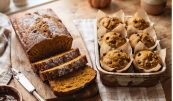 A rustic kitchen counter features a sliced loaf of pumpkin bread with melted chocolate chips alongside six parchment-lined muffins in a baking dish, a knife on a wooden board, a small bowl of chocolate spread, and a decorative pumpkin in the background.