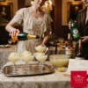 Two women in period-appropriate attire prepare a vintage-style Gilded Age lemon punch in an elegant, dining room setting, with one pouring champagne over a scoop of sorbet while the other holds a bottle of Canada Dry ginger ale next to bottles of Bacardi Gold rum and Courvoisier brandy and an open recipe book.
